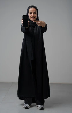 عرض شاشة سوداء فارغة عبر الهاتف المحمول، بورتريه لإمرأة عربية خليجية سعودية ترتدي الحجاب والعباءة السوداء تشير باصبع السبابة الى الجوال بتعابير السرور والسعادة، استخدام جهاز تقني حديث، الجسم بالطول الكامل، خلفية رمادية