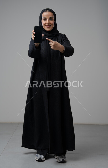 Blank black screen view on mobile phone, portrait of a Saudi Arabian Gulf woman wearing hijab and black abaya pointing index finger at mobile phone with expressions of pleasure and happiness, using modern technical device, full body, gray background