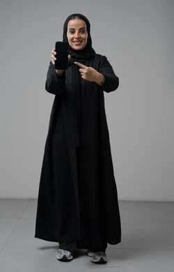 Blank black screen view on mobile phone, portrait of a Saudi Arabian Gulf woman wearing hijab and black abaya pointing index finger at mobile phone with expressions of pleasure and happiness, using modern technical device, full body, gray background