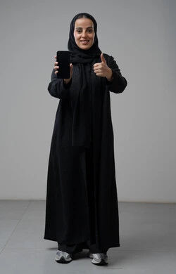 Blank black screen view on mobile phone, portrait of a Saudi Arabian Gulf woman wearing hijab and black abaya giving thumbs up with expressions of approval, approval and admiration, holding mobile phone in her hand, using modern technical device, full body, gray background