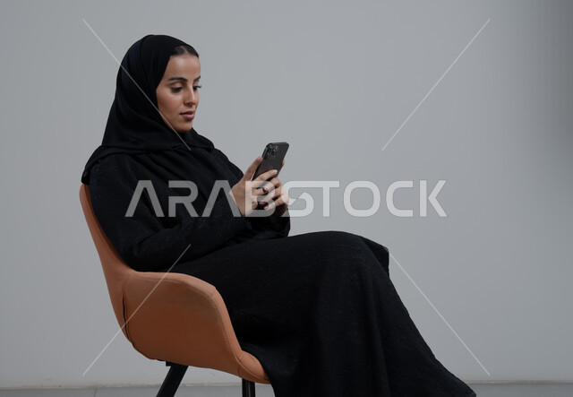 استخدام جهاز تقني حديث، بورتريه لإمرأة عربية خليجية سعودية ترتدي الحجاب والعباءة السوداء تجلس على كرسي مريح تقوم بالعمل على الهاتف المحمول بتعابير السرور والاندماج، خلفية رمادية