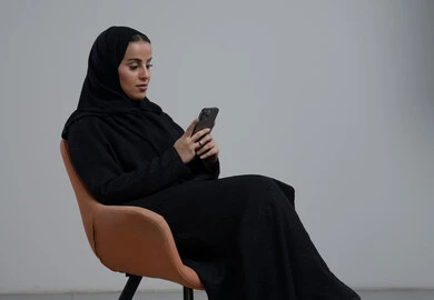 Using a modern technical device, portrait of a Saudi Arabian Gulf woman wearing a hijab and black abaya sitting on a comfortable chair working on a mobile phone with expressions of pleasure and integration, gray background