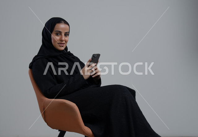 استخدام جهاز تقني حديث، بورتريه لإمرأة عربية خليجية سعودية ترتدي الحجاب والعباءة السوداء تجلس على كرسي مريح تقوم بالعمل على الهاتف المحمول بتعابير السرور والاندماج، خلفية رمادية