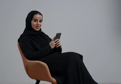 استخدام جهاز تقني حديث، بورتريه لإمرأة عربية خليجية سعودية ترتدي الحجاب والعباءة السوداء تجلس على كرسي مريح تقوم بالعمل على الهاتف المحمول بتعابير السرور والاندماج، خلفية رمادية