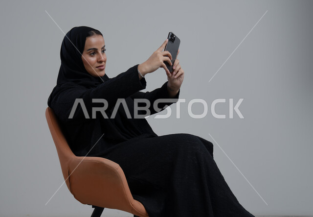 توثيق اللحظات الجميلة، التقاط صور سيلفي باستخدام الهاتف المحمول، بورتريه لإمرأة عربية خليجية سعودية ترتدي الحجاب والعباءة السوداء تجلس على كرسي تحمل بيدها الجوال بتعابير السرور والسعادة، استخدام جهاز تقني حديث، خلفية رمادية