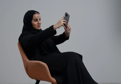 توثيق اللحظات الجميلة، التقاط صور سيلفي باستخدام الهاتف المحمول، بورتريه لإمرأة عربية خليجية سعودية ترتدي الحجاب والعباءة السوداء تجلس على كرسي تحمل بيدها الجوال بتعابير السرور والسعادة، استخدام جهاز تقني حديث، خلفية رمادية