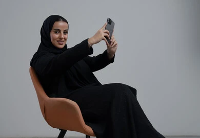 توثيق اللحظات الجميلة، التقاط صور سيلفي باستخدام الهاتف المحمول، بورتريه لإمرأة عربية خليجية سعودية ترتدي الحجاب والعباءة السوداء تجلس على كرسي تحمل بيدها الجوال بتعابير السرور والسعادة، استخدام جهاز تقني حديث، خلفية رمادية