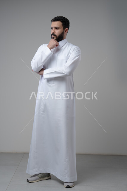 Looking forward and planning, problem solving and decision making, portrait of a Saudi Arabian Gulf man wearing a Saudi thobe, putting his hand on his chin with gestures of thinking and absentmindedness, concept of scrutiny and concentration, full body, gray background