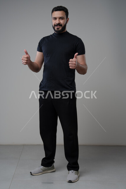 رفع اصابع الابهام الى الاعلى، النظر الى الكاميرا  بتعابير الاعجاب والرضا، بورتريه لرجل عربي خليجي سعودي يرتدي الزي الكاجوال ينظر الى الكاميرا بايماءات القبول والموافقة، صورة الجسم بالطول الكامل، خلفية رمادية