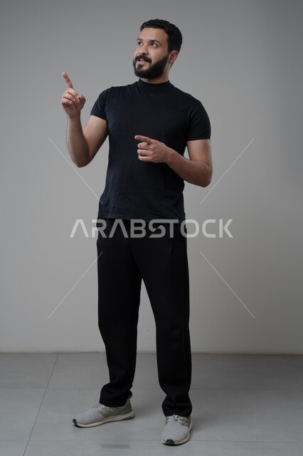 النظر الى الاعلى بتعابير التركيز، بورتريه لرجل عربي خليجي سعودي يرتدي الزي الكاجوال يشير بيده الى شيء ما، الدلالة على مكان معين، صورة الجسم بالطول الكامل، خلفية رمادية