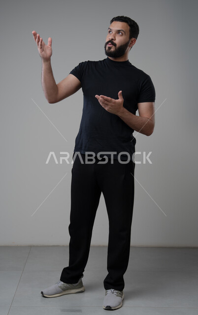 النظر الى الاعلى بتعابير التركيز، بورتريه لرجل عربي خليجي سعودي يرتدي الزي الكاجوال يشير بيده الى شيء ما، الدلالة على مكان معين، صورة الجسم بالطول الكامل، خلفية رمادية