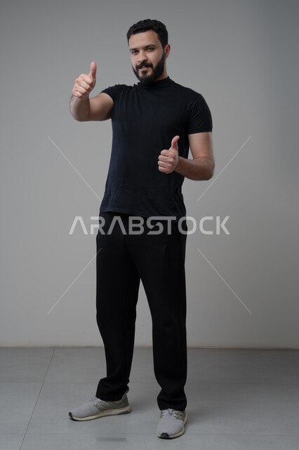 رفع اصابع الابهام الى الاعلى، النظر الى الكاميرا  بتعابير الاعجاب والرضا، بورتريه لرجل عربي خليجي سعودي يرتدي الزي الكاجوال ينظر الى الكاميرا بايماءات القبول والموافقة، صورة الجسم بالطول الكامل، خلفية رمادية