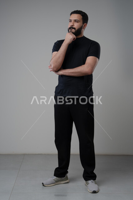 Looking forward and planning, problem solving and decision making, portrait of a Saudi Arabian Gulf man in casual wear putting his hand on his chin with gestures of thinking and absentmindedness, concept of scrutiny and concentration, full body, gray background
