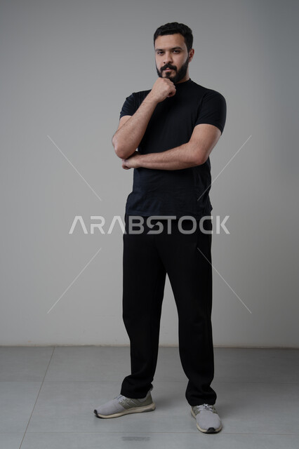 Looking forward and planning, problem solving and decision making, portrait of a Saudi Arabian Gulf man in casual wear putting his hand on his chin with gestures of thinking and absentmindedness, concept of scrutiny and concentration, full body, gray background