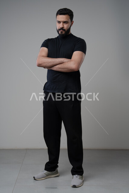  التقاط صور شخصية للوثائق الرسمية، الاعتناء والاهتمام بالمظهر الخارجي، مفهوم الاناقة والرجولة، بورتريه لشاب عربي خليجي سعودي يرتدي الزي الكاجوال يقف بأيدي متقاطعة ينظر الى الكاميرا بتعابير السرور والثقة بالنفس، الجسم بالطول الكامل، خلفية رمادية