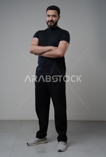  التقاط صور شخصية للوثائق الرسمية، الاعتناء والاهتمام بالمظهر الخارجي، مفهوم الاناقة والرجولة، بورتريه لشاب عربي خليجي سعودي يرتدي الزي الكاجوال يقف بأيدي متقاطعة ينظر الى الكاميرا بتعابير السرور والثقة بالنفس، الجسم بالطول الكامل، خلفية رمادية