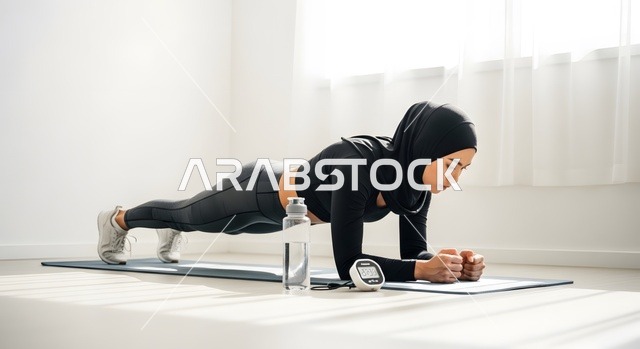 ضرورة شرب الماء اثناء الرياضة، القيام بالتمارين والأنشطة الجسديةوالبدنية، امرأة عربية خليجية سعودية محجبة ترتدي الملابس الرياضية تمارس تمرين البلانك على بساط رياضي، ممارسة تمارين رياضية عالية الكثافة في المنزل، أسلوب حياة صحي، الاهتمام بالصحة واللياقة