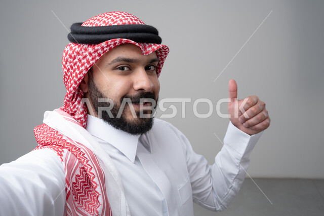 Thumbs up, looking at the camera with an expression of admiration and satisfaction, close-up portrait of a Saudi Arabian Gulf man wearing a traditional thobe and shemagh looking at the camera with gestures of acceptance and approval, taking a souvenir selfie, white background