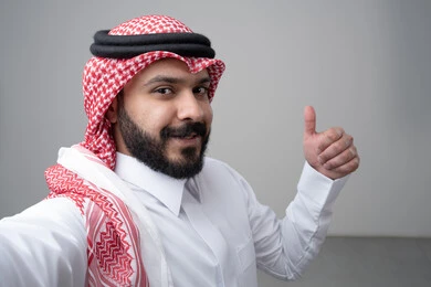 رفع اصابع الابهام الى الاعلى، النظر الى الكاميرا  بتعابير الاعجاب والرضا، بورتريه مقرب لرجل عربي خليجي سعودي يرتدي االثوب والشماغ التقليدي ينظر الى الكاميرا بايماءات القبول والموافقة، التقاط صورة سيلفي تذكارية، خلفية بيضاء