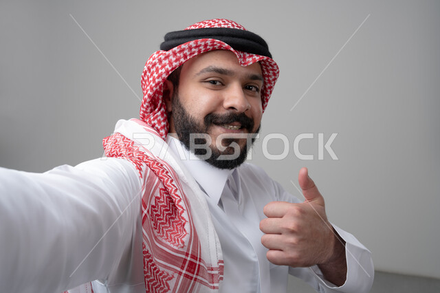 رفع اصابع الابهام الى الاعلى، النظر الى الكاميرا  بتعابير الاعجاب والرضا، بورتريه مقرب لرجل عربي خليجي سعودي يرتدي االثوب والشماغ التقليدي ينظر الى الكاميرا بايماءات القبول والموافقة، التقاط صورة سيلفي تذكارية، خلفية بيضاء