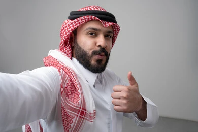 رفع اصابع الابهام الى الاعلى، النظر الى الكاميرا  بتعابير الاعجاب والرضا، بورتريه مقرب لرجل عربي خليجي سعودي يرتدي االثوب والشماغ التقليدي ينظر الى الكاميرا بايماءات القبول والموافقة، التقاط صورة سيلفي تذكارية، خلفية بيضاء