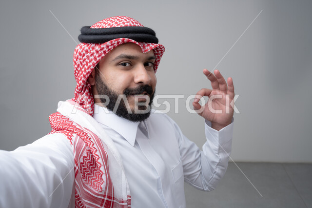 النظر الى الكاميرا  بتعابير الاعجاب والرضا، بورتريه مقرب لرجل عربي خليجي سعودي يرتدي االثوب والشماغ التقليدي يشير بيده بعلامة الامتياز، التقاط صورة سيلفي تذكارية، خلفية بيضاء