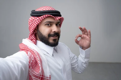 النظر الى الكاميرا  بتعابير الاعجاب والرضا، بورتريه مقرب لرجل عربي خليجي سعودي يرتدي االثوب والشماغ التقليدي يشير بيده بعلامة الامتياز، التقاط صورة سيلفي تذكارية، خلفية بيضاء