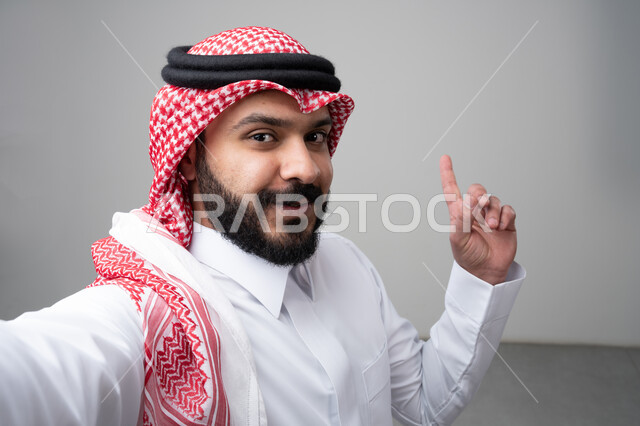  الإشارة بإصبع السبابة إلى شيء ما، بورتريه مقرب لرجل عربي خليجي سعودي يرتدي االثوب والشماغ التقليدي ينظر إلى الكاميرا بإيماءات السعادة والسرور، التقاط صورة سيلفي تذكارية، خلفية بيضاء