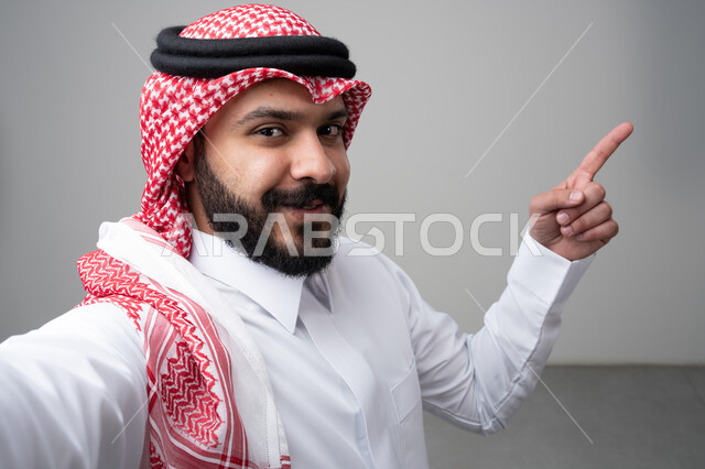  الإشارة بإصبع السبابة إلى شيء ما، بورتريه مقرب لرجل عربي خليجي سعودي يرتدي االثوب والشماغ التقليدي ينظر إلى الكاميرا بإيماءات السعادة والسرور، التقاط صورة سيلفي تذكارية، خلفية بيضاء