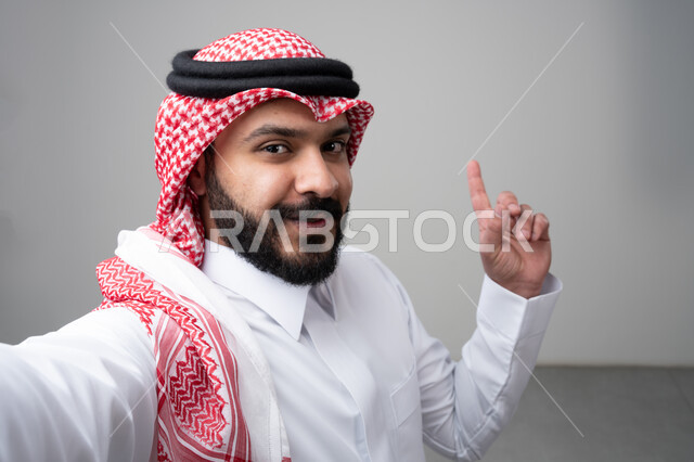  الإشارة بإصبع السبابة إلى شيء ما، بورتريه مقرب لرجل عربي خليجي سعودي يرتدي االثوب والشماغ التقليدي ينظر إلى الكاميرا بإيماءات السعادة والسرور، التقاط صورة سيلفي تذكارية، خلفية بيضاء