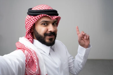  الإشارة بإصبع السبابة إلى شيء ما، بورتريه مقرب لرجل عربي خليجي سعودي يرتدي االثوب والشماغ التقليدي ينظر إلى الكاميرا بإيماءات السعادة والسرور، التقاط صورة سيلفي تذكارية، خلفية بيضاء