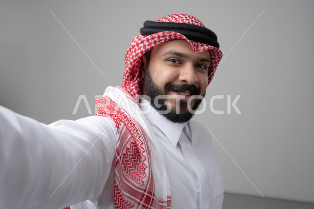 Front camera shot, close-up portrait of a Saudi Arabian Gulf man wearing a traditional thobe and shemagh looking at the camera with happy gestures, taking a souvenir selfie, white background