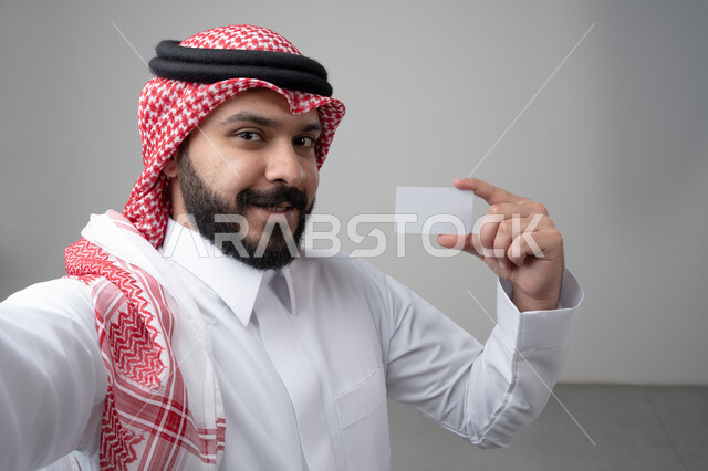 عرض بطاقة عمل تعريفية شخصية، الشراء والدفع بطريقة بطاقات الائتمان البنكية، بورتريه مقرب لرجل عربي خليجي سعودي يرتدي االثوب والشماغ التقليدي يمسك بطاقة بيضاء فارغة، النظر إلى الكاميرا بإيماءات السعادة والسرور، التقاط صورة سيلفي تذكارية، خلفية بيضاء