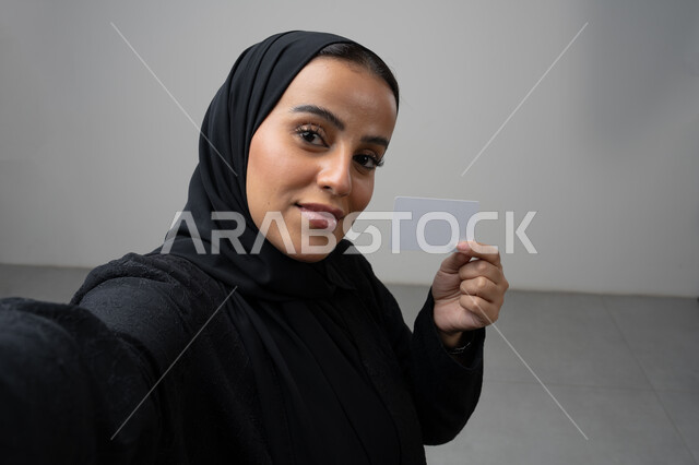 عرض بطاقة عمل تعريفية شخصية، الشراء والدفع بطريقة بطاقات الائتمان البنكية، بورتريه مقرب لامرأة عربية خليجية سعودية ترتدي الحجاب والعباءة السوداء تمسك بطاقة بيضاء فارغة، النظر إلى الكاميرا بإيماءات السعادة والسرور، التقاط صورة سيلفي تذكارية، خلفية بيضاء