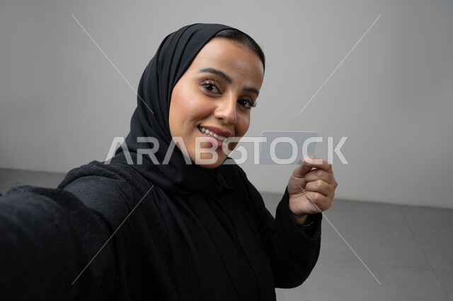 عرض بطاقة عمل تعريفية شخصية، الشراء والدفع بطريقة بطاقات الائتمان البنكية، بورتريه مقرب لامرأة عربية خليجية سعودية ترتدي الحجاب والعباءة السوداء تمسك بطاقة بيضاء فارغة، النظر إلى الكاميرا بإيماءات السعادة والسرور، التقاط صورة سيلفي تذكارية، خلفية بيضاء