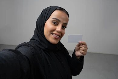 عرض بطاقة عمل تعريفية شخصية، الشراء والدفع بطريقة بطاقات الائتمان البنكية، بورتريه مقرب لامرأة عربية خليجية سعودية ترتدي الحجاب والعباءة السوداء تمسك بطاقة بيضاء فارغة، النظر إلى الكاميرا بإيماءات السعادة والسرور، التقاط صورة سيلفي تذكارية، خلفية بيضاء