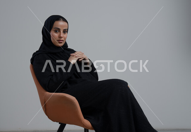 الاهتمام بالأناقة والمظهر الخارجي، تعابير وإيماءات الثقة بالنفس, بورتريه لشابة عربية خليجية سعودية محجبة تجلس على كرسي مريح بأيدي متشابكة ترتدي العباءة السوداء, مفهوم النعومة والأنوثة، العناية بالجمال والشياكة، خلفية بيضاء