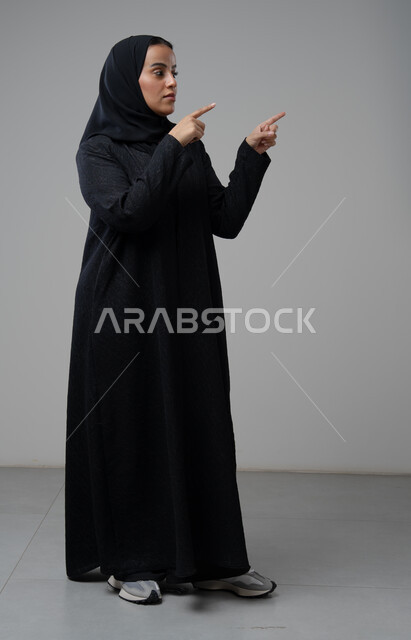 الإشارة إلى عنصر ما, التوضيح والدلالة لمكان محدد, بورتريه لامرأة عربية خليجية سعودية محجبة ترتدي العباءة السوداء تشير بيديها على الجانب، تحديد موقع شيء معين, صورة الجسم بالطول الكامل، خلفية بيضاء