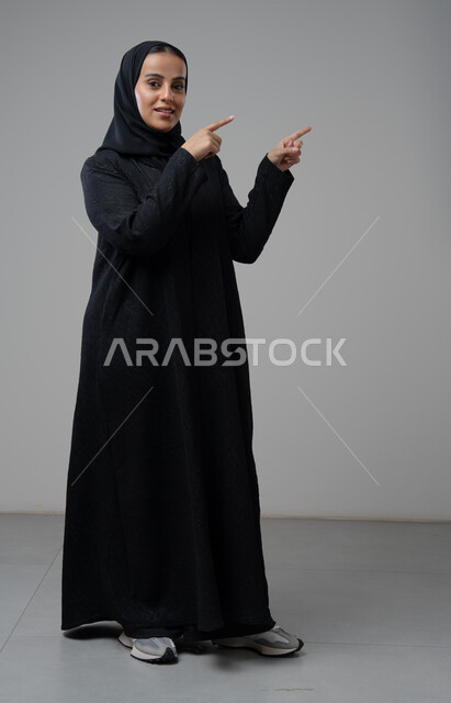 الإشارة إلى عنصر ما, التوضيح والدلالة لمكان محدد, بورتريه لامرأة عربية خليجية سعودية محجبة ترتدي العباءة السوداء تشير بيديها على الجانب، تحديد موقع شيء معين, صورة الجسم بالطول الكامل، خلفية بيضاء