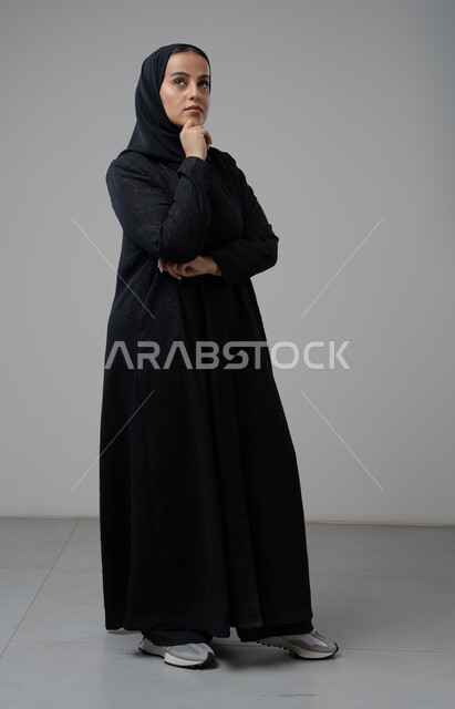 Gestures of thinking and contemplating something, portrait of a veiled Saudi Arabian Gulf woman wearing a black abaya, putting her hand on her face with gestures of contemplation, full-length body image, white background