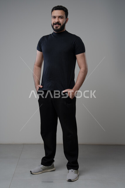  الاعتناء والاهتمام بالمظهر الخارجي، ايماءات السعادة والسرور، مفهوم الاناقة والرجولة، بورتريه لشاب عربي خليجي سعودي يرتدي الزي الكاجوال يقف باستقامة ينظر الى الكاميرا بتعابير الثقة بالنفس، الجسم بالطول الكامل، خلفية رمادية