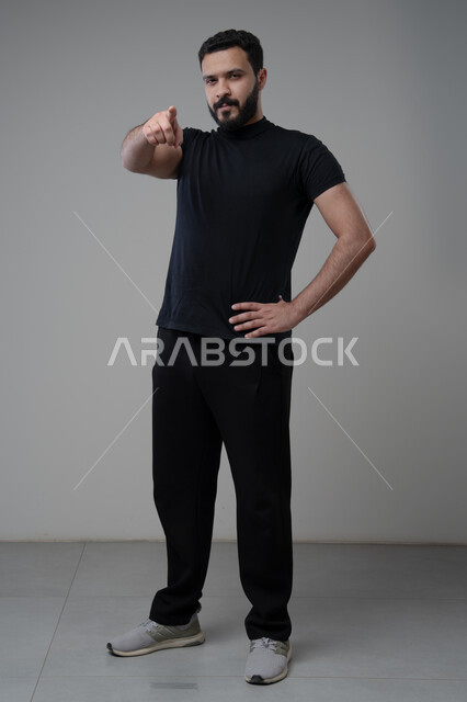 الاشارة إلى شيء ما بإيماءات السرور ,تعابير التأكد والتحديد,  بورتريه لرجل عربي خليجي سعودي مبتسم يرتدي الزي الكاجوال يرفع إصبعه السبابة للاختيار وينظر إلى الكاميرا بتعابير الجدية والحزم, صورة للجسم بالكامل, خلفية رمادية