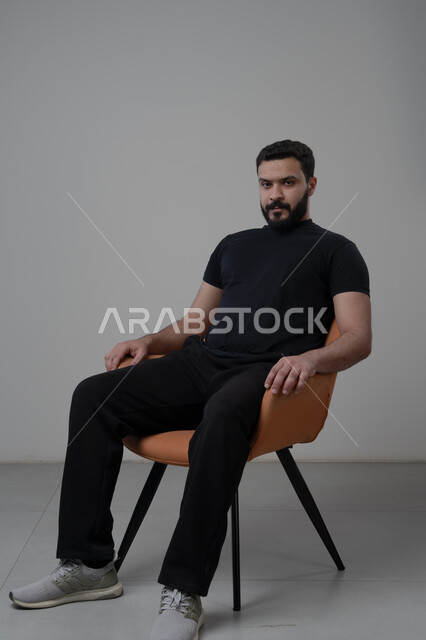  الاعتناء والاهتمام بالمظهر الخارجي، الجلوس بإيماءات الجدية والحزم، مفهوم الاناقة والرجولة، بورتريه لشاب عربي خليجي سعودي يرتدي الزي الكاجوال يجلس على كرسي وينظر الى الكاميرا بتعابير الثقة بالنفس، خلفية بيضاء