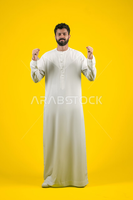 The concept of patience and deliberation, calmness and request to wait, practicing relaxation exercises, a portrait of an Arab Gulf Emirati man wearing a white kandura raising his hands in gestures of patience, giving time to do something, a full body image, a yellow background