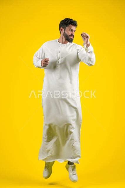 التدرب على إتقان فنون الملاكمة، زيادة قدرة العضلات على التحمل، بورتريه لشاب عربي خليجي اماراتي يرتدي الكندورة يقفز عاليا ويرفع قبضات يديه بإيماءات القوة، ممارسة رياضة الدفاع عن النفس، الجسم بالطول الكامل، خلفية صفراء