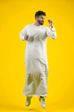 التدرب على إتقان فنون الملاكمة، زيادة قدرة العضلات على التحمل، بورتريه لشاب عربي خليجي اماراتي يرتدي الكندورة يقفز عاليا ويرفع قبضات يديه بإيماءات القوة، ممارسة رياضة الدفاع عن النفس، الجسم بالطول الكامل، خلفية صفراء