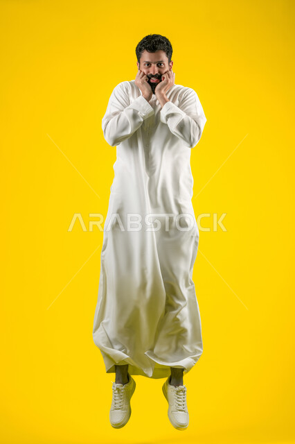 Receiving unexpected happy news, a young Emirati man looking at the camera with gestures of shock, amazement and amazement, being surprised by a specific situation, feeling astonished and amazed, remembering something, portrait of a Saudi Arabian Gulf man wearing a kandura and putting his hands on his face, full body photo, yellow background