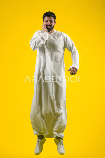 Tooth decay and gingivitis, toothache and pain from cavities, need to go to the dental clinic, feeling tired and sick, portrait of a young Emirati Gulf Arab man wearing a kandura jumping high and putting his hands on his cheeks with an expression of pain, full body, yellow background