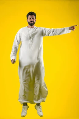 The concept of elegance and interest in outward appearance, a portrait of a young Arab Gulf Emirati man wearing a kandura, standing straight and looking at the camera with self-confident gestures, pointing with his index finger at something, full-length body, yellow background