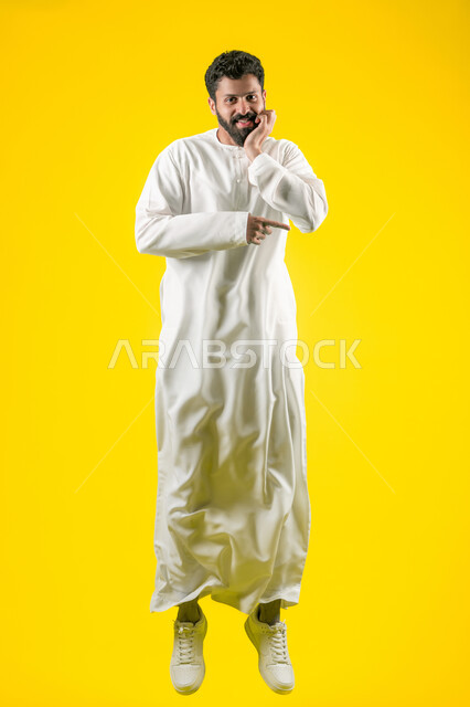 Gestures of boredom and disappointment, the concept of elegance and interest in outward appearance, a portrait of a young Emirati Gulf Arab man wearing a kandura, jumping high, looking at the camera, putting his hand on his face and pointing with his index finger at something, full body, yellow background